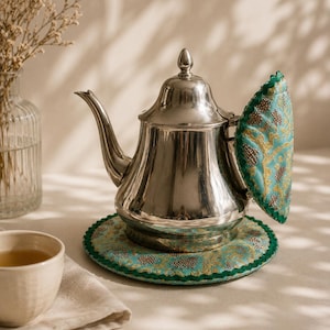 May include: A polished silver-toned metal teapot featuring a hinged lid and a curved spout. The teapot is accompanied by a circular, padded fabric base and a matching semi-circular handle cover, both in a teal fabric with gold embroidery.