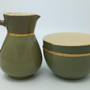 May include: A ceramic creamer jug and a matching sugar bowl, both featuring a smooth olive green exterior, a cream-coloured interior, and a decorative gold-toned band encircling the middle of each piece with a textured rope-like pattern.