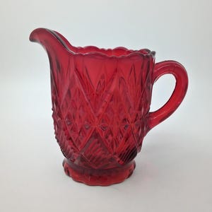 May include: A deep red glass creamer jug featuring a prominent diamond-cut pattern across its body. The vessel has a scalloped rim, a curved pouring spout, and a sturdy handle on the side, all crafted from translucent, vibrant ruby-coloured glass.