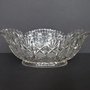 May include: A clear glass bowl featuring an intricate pressed pattern with diamond-shaped cross-hatching and vertical fan-like motifs. The bowl has a scalloped, serrated rim and sits on a smooth, oval-shaped base, showcasing a reflective surface.