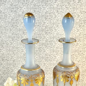 May include: Two frosted glass perfume bottles with light blue stoppers and gold accents. The bottles have a bulbous shape with gold filigree designs. The stoppers are light blue with gold trim.