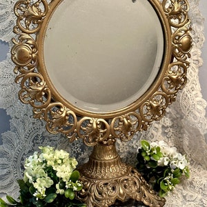 May include: Ornate gold-colored vanity mirror with a round, beveled glass. The mirror is supported by a decorative pedestal base and features an elaborate, scrolling floral design around the frame. The mirror is approximately 12 inches in diameter.
