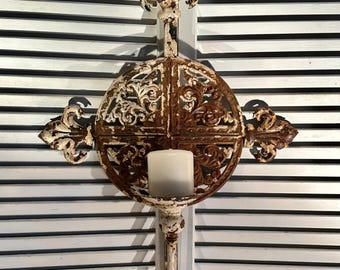 May include: A wall-mounted metal candle holder featuring a distressed white and rust-colored finish. The design includes a central circular frame with intricate scrollwork, fleur-de-lis accents at the top, sides, and bottom, and a small platform.
