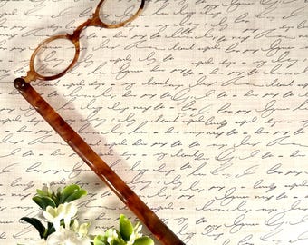 May include: A lorgnette with a brown tortoiseshell pattern handle and matching circular frames. The handle is long and slender, featuring a small loop at the bottom. The frames are connected to the top of the handle by a hinge mechanism.