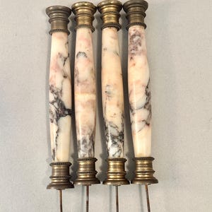 May include: Four antique marble and brass furniture pulls. The pulls have a cylindrical shape with a marble body featuring pink and gray veining. The top and bottom are brass with a decorative design. Each pull has a metal rod extending from the bottom.