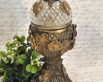 May include: A decorative glass and brass container featuring a diamond-cut glass lid topped with a brass bee. The base is encased in a brass cage adorned with metal flowers and leaves, creating a structured, ornate vessel for small items.