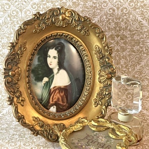 May include: A gold-toned oval frame with a portrait of a woman in a green and red dress.  The frame is sitting on a table next to a gold-toned ring box with the word "Rings" on the top and a clear glass bottle with a gold-toned top.
