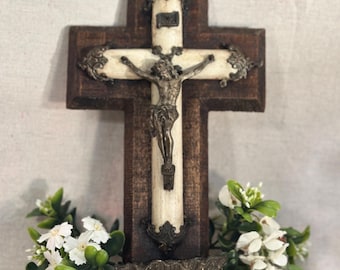 May include: A wall-mounted crucifix featuring a dark brown wooden cross base with a central off-white vertical inlay. A metallic silver-toned figure of Jesus is attached to the center, accented by ornate metal filigree at the ends and a small shell-shaped basin at the bottom.