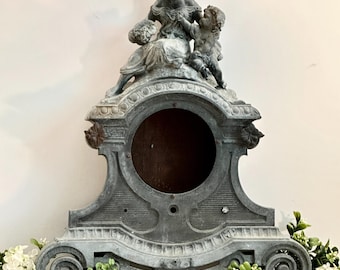May include: A gray metal clock housing featuring a sculptural topper of a seated figure and a small child. The frame has an ornate, arched design with a large circular opening in the center and decorative scrollwork at the base of the structure.