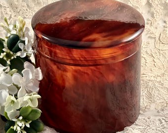 May include: A cylindrical lidded container featuring a polished, marbled finish in deep shades of amber, brown, and black. The surface displays a swirling, translucent pattern throughout the body and the circular lid, creating a glossy appearance.