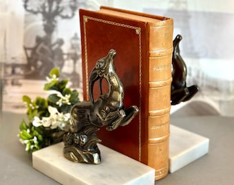 May include: A pair of bookends featuring bronze-toned metal gazelles leaping over textured bases, each mounted on a rectangular white marble slab. The bookends support a vintage leather-bound book with gold-embossed lettering on the spine.