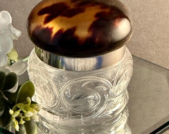 May include: A small glass jar with a clear, embossed floral pattern on the body and a smooth, rounded lid featuring a brown and black tortoiseshell pattern. The lid is secured by a polished silver-toned metal band that encircles the top of the jar.