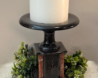 May include: A white cylindrical pillar candle sits atop a black and reddish-brown marble pedestal candle holder. The base features a square design with inlaid stone panels and delicate gold-toned line art detailing on the front face.
