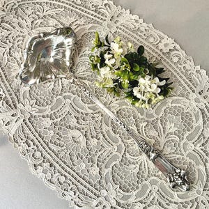 May include: A silver ladle with a decorative handle and a leaf-shaped bowl. The ladle is placed on a white lace tablecloth, with a small bouquet of white flowers and green leaves nearby. The ladle is likely vintage or antique.