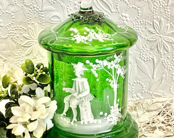 May include: A cylindrical green glass jar with a matching lid featuring a clear glass finial. The jar is decorated with white painted details depicting a person sitting on a stool next to a tree, with additional white floral accents on the lid.