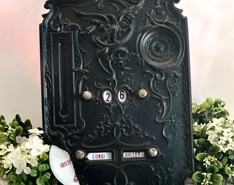 May include: A black wall-mounted perpetual calendar featuring ornate relief carvings of an eagle and floral motifs. The calendar displays the number 26 and the French text LUNDI JUILLET, with a small white circular disc showing the word JEUDI.