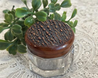 May include: A small, clear, hexagonal glass jar with a round, dark brown lid. The lid features a textured, raised, copper-colored surface that resembles tree bark. The jar is empty and sits upright, showcasing its multi-faceted, transparent base.