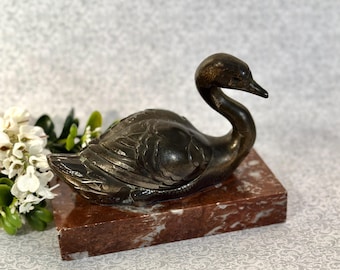May include: A dark bronze-toned metal swan figurine with detailed wing carvings, mounted on a rectangular base made of polished reddish-brown marble with white veining. The swan is depicted in a resting position with its neck curved forward.