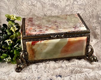 May include: A rectangular box crafted from polished green and orange marbled stone, featuring a hinged lid and ornate metallic trim. The box is supported by four decorative metal feet shaped like stylized fish, providing a stable base.