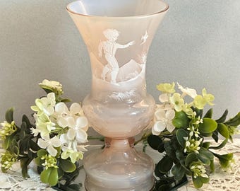 May include: A pale pink opalescent glass vase featuring a flared rim and a pedestal base. The exterior surface is decorated with an etched white relief design depicting a figure walking through a landscape with mountains and a bird in flight.