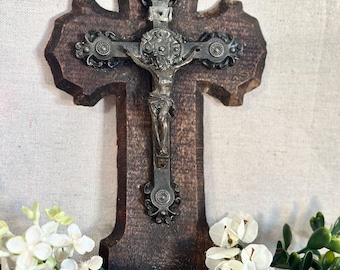 May include: A dark brown wooden wall cross featuring a metallic silver-toned crucifix figure at the center. The cross has ornate decorative metal accents at the top, sides, and bottom, with a small basin attached to the base of the wooden structure.