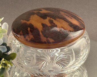 May include: A clear glass jar featuring intricate etched floral patterns on its rounded body, topped with a smooth, polished lid displaying a mottled brown and black tortoiseshell-like pattern. The container has a wide, circular base.