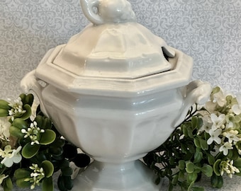 May include: A white ceramic tureen with an octagonal base, a matching lid featuring a sculpted handle, and two side handles. The vessel has a smooth, glossy finish and a faceted body design, providing a classic look for serving soups or stews.