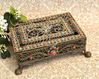 May include: A rectangular metal filigree box featuring intricate scrollwork, four small ball feet, and a central cherub motif on the lid. The box is decorated with alternating small round turquoise and coral-colored cabochons along the edges.