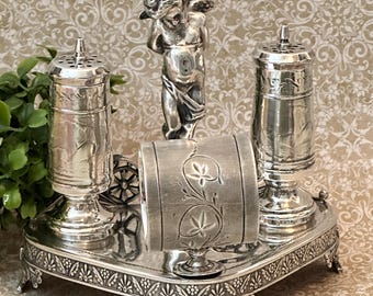 May include: A silver-toned condiment set featuring two cylindrical shakers with perforated tops, a central cherub figurine, and a napkin ring with floral engravings, all mounted on a rectangular base with ornate decorative feet and edges.