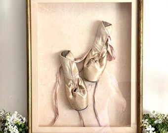 May include: A pair of pale pink satin ballet slippers with matching ribbons, displayed inside a rectangular gold-toned frame. The slippers are positioned against a soft pink background, creating a shadow box effect with a clear glass front.