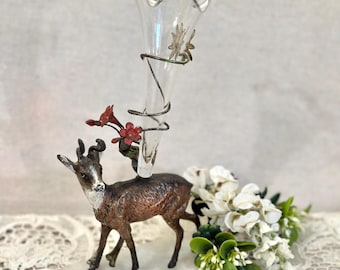May include: A brown metal deer figurine standing on a rectangular white marble base. A clear glass trumpet-shaped vase is attached to the deer's back, wrapped in a metal vine with small red flowers and a silver star-shaped accent piece.