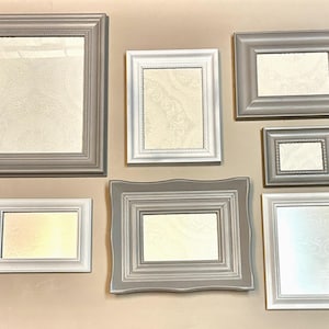 May include: A collection of seven picture frames in various sizes and shapes. The frames are painted in shades of gray and white, with a decorative pattern visible inside the frames. The frames are mounted on a neutral-colored wall.