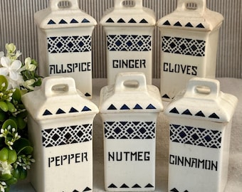 May include: A set of six off-white ceramic spice jars with dark blue geometric patterns and handles on the lids. The jars are labeled Allspice, Ginger, Cloves, Pepper, Nutmeg, and Cinnamon in black lettering. Each jar has a rectangular body shape.