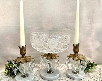 May include: Two brass and marble candle holders with hanging crystal prisms, each holding a tall white taper candle, and a matching crystal bowl with floral etching on a central marble base, all featuring ornate metal petal-shaped accents.