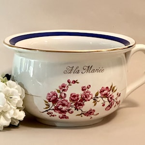 May include: A white ceramic chamber pot with a blue and gold rim. The pot has a floral design and the words "A la Mariée" written on the side.