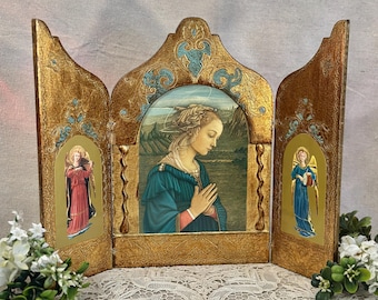 May include: A triptych featuring a central panel with a woman in a blue dress with hands clasped in prayer, flanked by two side panels depicting angels in red and blue robes. The frame is gold with ornate carvings and blue accents.