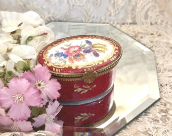 May include: An oval-shaped trinket box with a deep red base and a hinged lid. The lid features a white center panel decorated with a colorful floral bouquet, surrounded by a gold-toned ornate border. The box has a metallic rim and hinge.