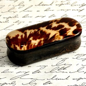 May include: A small, rectangular box with rounded corners featuring a polished lid with a mottled brown and cream pattern. The base of the box is a solid, dark brown color with a smooth, matte finish, contrasting with the glossy, patterned lid.