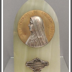 May include: A religious plaque featuring a gold-toned medallion with a profile of a woman in a veil, inscribed with French text. The plaque is set on a light green, arched base with a silver-toned inscription below.