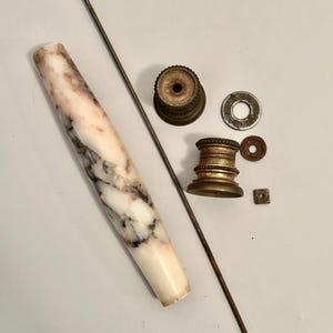 K&ouml;nnte beinhalten: Eine Ansammlung antiker Hardware-Komponenten, darunter ein Griff mit Marmormuster, Messingbeschl&auml;ge, Unterlegscheiben und eine Gewindestange. Der Griff ist wei&szlig; mit rosa und grauen Adern. Die Messingteile weisen Altersspuren auf.