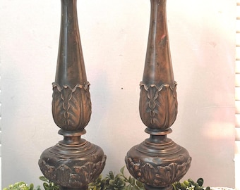 May include: Two bronze-colored candlestick holders. Each has a round base with leaf detailing, a bulbous midsection with more leaf designs, and a tall, slender stem. The top has a small, rounded cup for holding a candle. The overall design is ornate and vintage.