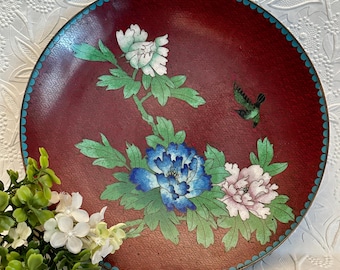 May include: A round cloisonné plate with a deep red background featuring a textured pattern. The plate displays a floral design with white, blue, and pink peony blossoms, green leaves, and a small green bird. The rim is accented with a blue border.