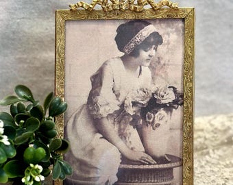 May include: A rectangular gold metal picture frame with an ornate embossed border and a decorative bow crest at the top. The frame contains a sepia-toned photograph of a person wearing a headband and dress, leaning over a vase of flowers.