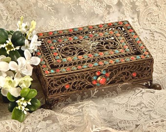 May include: A rectangular metal box with an intricate filigree design, featuring a central cherub motif on the lid. The box is adorned with small, alternating red and turquoise circular cabochons along the edges and a floral-shaped clasp.