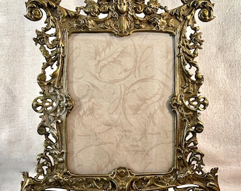 May include: A rectangular gold-toned metal picture frame featuring ornate scrollwork, floral motifs, and a central human face relief at the top. The frame has a wide, decorative border and stands on two curved feet, enclosing a light beige insert.