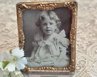 May include: A rectangular gold-toned metal frame with an ornate, embossed floral border containing a black and white vintage photograph of a young child with curly hair wearing a collared dress. The frame is supported by a clear acrylic stand.