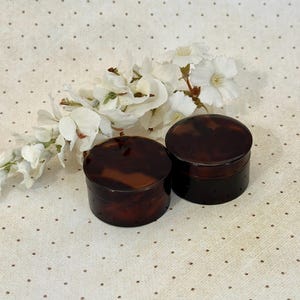 May include: Two small, round, lidded containers featuring a glossy, dark brown and black tortoiseshell pattern. The containers are positioned side-by-side, with a cluster of white flowers resting behind them, adding a natural element to the composition.