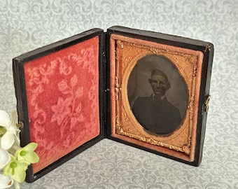 May include: A hinged, dark-colored case containing a black and white portrait of a person in a suit. The interior features a red velvet lining with a floral pattern on the left and an ornate, oval-shaped gold-toned metal frame on the right.