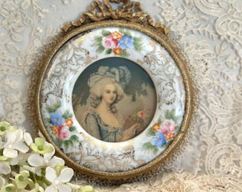 May include: A circular porcelain wall plaque featuring a central portrait of a person in 18th-century attire, surrounded by a white border with hand-painted floral motifs in pink, blue, and green, encased in an ornate, antiqued gold metal frame.