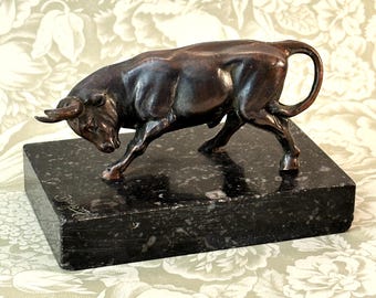 May include: A bronze-toned bull figurine in a charging stance, featuring a muscular build and curved horns. The bull is mounted on a rectangular, polished black marble base with visible natural speckling and an etched signature on the side.